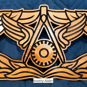 May include: A wooden plaque with a black outline of a military badge. The badge features a compass and a gear, surrounded by wings. The plaque is labeled "Knotty Alder".