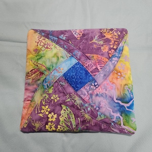 May include: A colorful, quilted square with a patchwork design. The fabric features a vibrant mix of purple, yellow, blue, and green hues. The design includes floral and abstract patterns, with a central blue square. The edges are neatly stitched.
