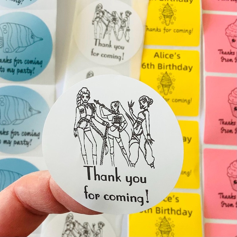 Pode incluir: Um adesivo redondo branco com um desenho de linha preta de tr&ecirc;s personagens femininas em equipamento t&aacute;tico, com o texto Thank you for coming! impresso abaixo. O adesivo faz parte de uma folha com outros r&oacute;tulos azuis, amarelos e rosas.