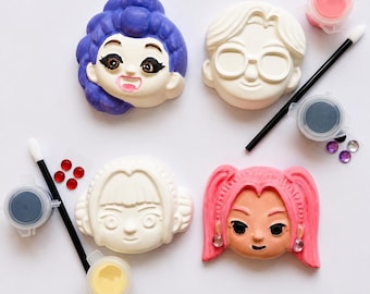 May include: A DIY craft kit featuring four sculpted character faces, two black paintbrushes, four small paint pots in pink, black, yellow, and white, and several small adhesive rhinestones in red, purple, and clear colors.