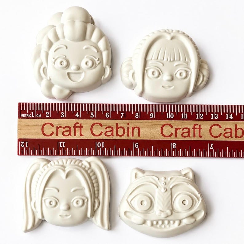 Pode incluir: Quatro moldes de silicone em tom esbranqui&ccedil;ado com rostos de personagens estilizados, incluindo tr&ecirc;s figuras humanas com penteados distintos e um rosto animal. Uma r&eacute;gua de madeira com o texto Craft Cabin est&aacute; colocada no centro.