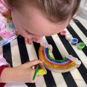 May include: A child paints a wooden rainbow with colourful paint. The rainbow has a layered effect with each colour showing through the next. The rainbow is on a black and white striped surface.