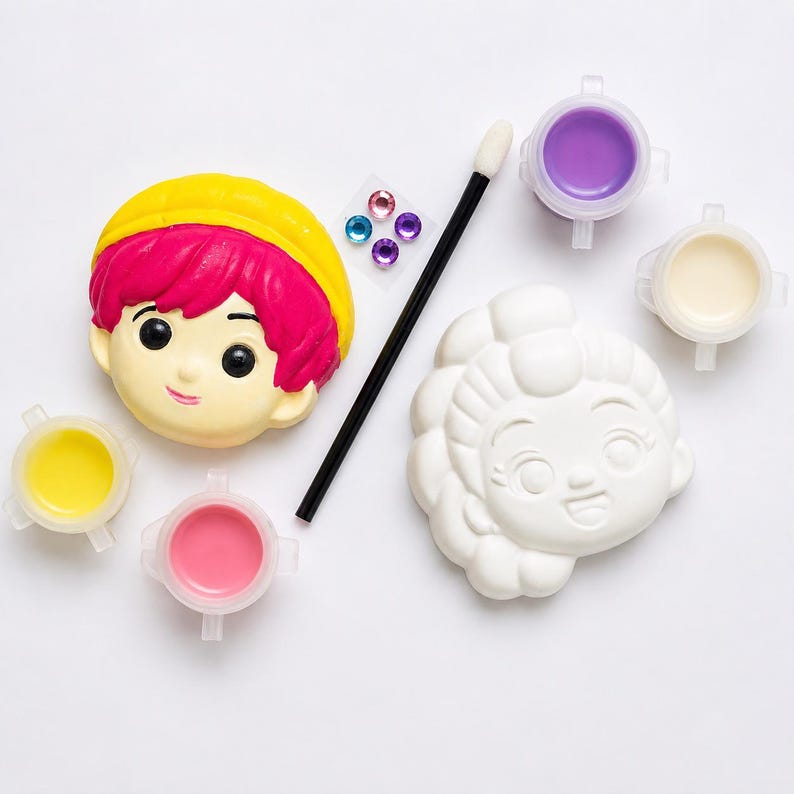 Pode incluir: Um kit de artesanato com uma cabe&ccedil;a de personagem pintada com cabelo rosa brilhante e chap&eacute;u amarelo, uma cabe&ccedil;a branca sem pintura, um pincel preto, quatro potes de tinta em amarelo, rosa, roxo e creme, e quatro pequenas gemas adesivas.