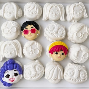 Pode incluir: Uma cole&ccedil;&atilde;o de dezasseis pequenas figuras em forma de cabe&ccedil;a de personagem dispostas numa grelha quatro por quatro. A maioria &eacute; de cor branca lisa, enquanto tr&ecirc;s apresentam detalhes pintados, incluindo cabelo preto com &oacute;culos cor-de-rosa, cabelo cor-de-rosa brilhante com uma fita amarela e cabelo roxo.
