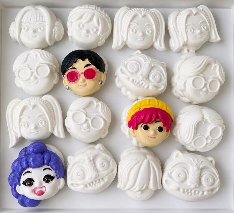 Pode incluir: Uma cole&ccedil;&atilde;o de dezasseis pequenas figuras em forma de cabe&ccedil;a de personagem dispostas numa grelha quatro por quatro. A maioria &eacute; de cor branca lisa, enquanto tr&ecirc;s apresentam detalhes pintados, incluindo cabelo preto com &oacute;culos cor-de-rosa, cabelo cor-de-rosa brilhante com uma fita amarela e cabelo roxo.