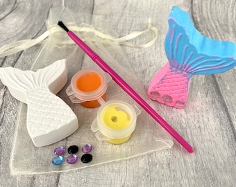 May include: A craft kit for painting mermaid tails. The kit includes two white ceramic mermaid tails, two small bottles of paint, a paintbrush, and a bag of rhinestones.