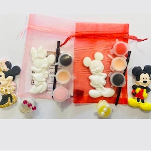 Può includere: Due piccole borse rosa con coulisse con statuette di Topolino e Minnie da dipingere. Ogni borsa contiene una statuetta, vasetti di vernice e un pennello. Sono visibili anche altre statuette di Topolino e Minnie.