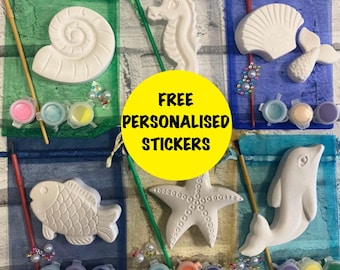 May include: A craft kit featuring white plaster sea-themed figures including a shell, seahorse, fish, starfish, and dolphin. Each set includes a paintbrush, small paint pots, and a sheer organza bag. A yellow circle displays the text FREE PERSONALISED STICKERS.