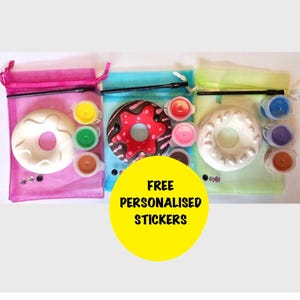 May include: Three paint-your-own donut kits in pink, blue, and green mesh bags. Each kit includes a ceramic donut, paint pots, and a paintbrush. A yellow circle with the text "FREE PERSONALISED STICKERS" is in the foreground.