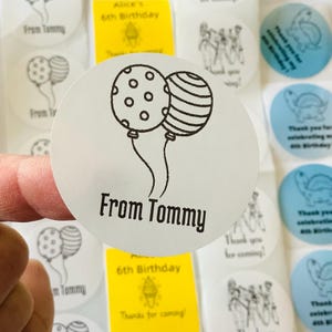 Pode incluir: Um autocolante circular branco com um desenho de linha preta de dois bal&otilde;es, um com padr&atilde;o de bolinhas e outro com riscas horizontais, com o texto From Tommy impresso numa fonte preta a negrito por baixo dos bal&otilde;es.