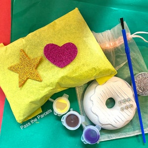May include: A craft kit for decorating a white donut-shaped ceramic with paint and glitter. The kit includes a paintbrush, three small paint pots, a small container of glitter, and a yellow tissue paper bag with a pink heart and gold star.