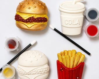 May include: A DIY craft kit featuring unpainted and painted ceramic figures of a burger, a cup with the word COLA, and french fries. The set includes two black paintbrushes with white tips and three small circular paint pots in red, yellow, and black.