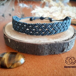 May include: A dark gray macrame bracelet with a woven pattern, displayed on a wooden slice. The bracelet is adjustable and sits atop a natural wood coaster. A polished tiger's eye stone and the logo "MACRA'ADDICT" are also visible.