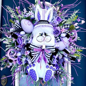 May include: A purple and white Easter wreath with a bunny wearing a top hat and glasses. The wreath is decorated with purple flowers, ribbon, and eggs.