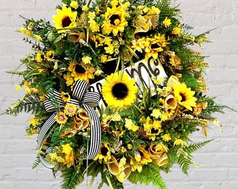 May include: A sunflower wreath with a welcome sign. The wreath is made of artificial sunflowers, greenery, and a black and white striped ribbon.
