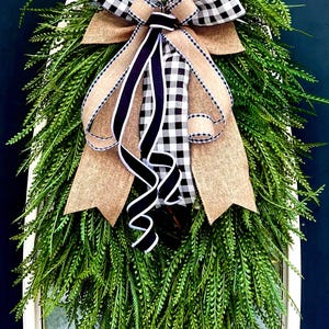May include: A teardrop-shaped door swag featuring lush green fern-like foliage. The arrangement is accented with a large decorative bow made of black and white buffalo check fabric, natural tan burlap, and solid black ribbon with white edges.