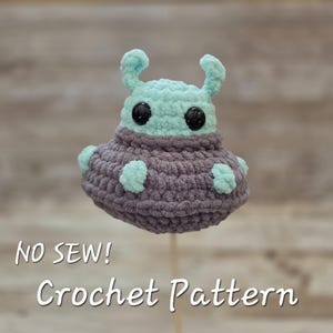 May include: A handmade crochet alien toy in shades of teal and gray. The alien has large black eyes and small teal appendages. The text "NO SEW! Crochet Pattern" is displayed in white script. The toy is mounted on a wooden stick.
