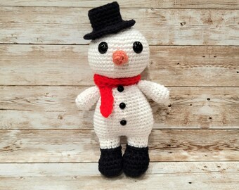 May include: A white crocheted snowman with a black top hat, red scarf, and black button eyes. The snowman is standing on a light brown wooden surface.