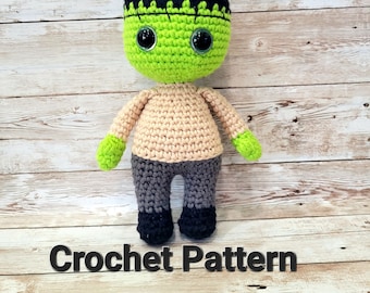 Könnte beinhalten: Gehäkelte Frankenstein-Puppe mit grüner Haut, schwarzen Haaren und grauen Hosen. Die Puppe hat große schwarze Augen und eine genähte Narbe auf der Stirn. Die Puppe sitzt auf einer Holzoberfläche. Häkelanleitung.