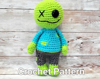 Könnte beinhalten: Gehäkelte grüne und blaue Zombie-Puppe mit einem Auge und einem genähten Mund. Die Puppe trägt eine graue Hose und hat ein "Crochet Pattern"-Textetikett darunter.
