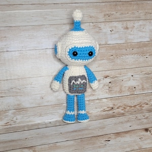 Puede incluir: Un juguete robot de crochet con un cuerpo blanco, brazos y piernas azules y una cara azul con ojos negros. El robot tiene un panel gris en el pecho con un diseño de línea ondulada.
