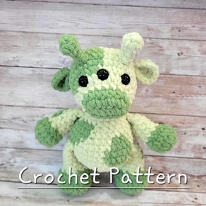 May include: A handmade crochet cow with a green and white spotted pattern. The plush toy has three black button eyes and small horns. The text "Crochet Pattern" is displayed at the bottom of the image.