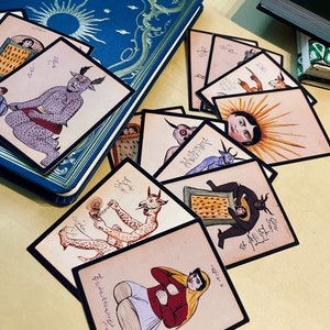 May include: A set of vintage playing cards featuring colourful illustrations of mythical creatures and figures. The cards are printed on thick paper with black borders and have Arabic text on them. The cards are arranged in a fan shape on a light brown surface.