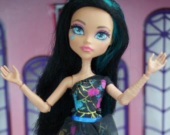 May include: A fashion doll with black hair and teal streaks, dressed in a black dress with a floral design and a blue belt. The doll has blue eyes and stands on a black stand.
