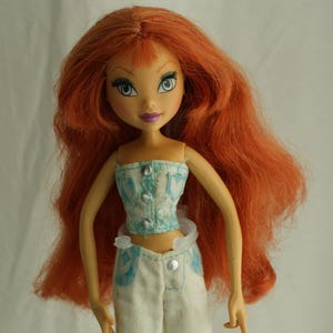 Puede incluir: Muñeca de moda con pelo largo y rojo vibrante y ojos azules. La muñeca lleva un top corto turquesa y blanco con botones plateados, pantalones blancos con patrones de estrellas azules y sandalias azules. La muñeca está sobre un soporte de plástico transparente.