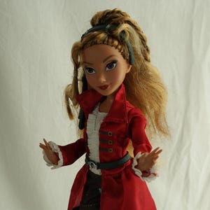 Könnte beinhalten: Puppe in einem roten Mantel im Piratenstil mit schwarzem Besatz, einem weißen Rüschenhemd und schwarzen Shorts. Die Puppe hat lange blonde Haare und trägt schwarze Stiefel. Eine kleine Schatztruhe steht am Fuß des Ständers.