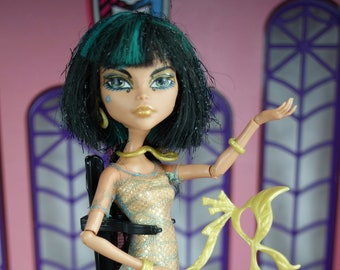 May include: A fashion doll with green hair and a gold and blue dress. She is holding a gold mask and standing on a black base. The doll is in front of a pink and purple background.
