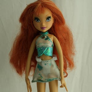 Könnte beinhalten: Puppe mit langen, roten Haaren und blauen Augen, bekleidet mit einem türkis-weißen Outfit. Die Puppe hat einen hellen Teint und steht auf einer transparenten Kunststoffbasis. Das Outfit umfasst ein Neckholder-Top und einen Rock.