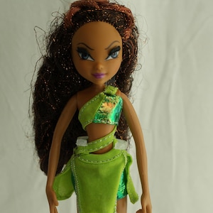 Może przedstawiać: Lalka modowa z brązowymi włosami i niebieskimi oczami, ubrana w zielony i opalizujący top i spódnicę. Lalka ma opaloną cerę i stoi na przezroczystym plastikowym stojaku. Strój jest ozdobiony białymi szwami.
