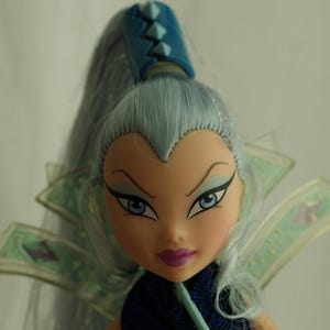 May include: A doll with pale skin, blue eyes, and light blue hair styled in a high ponytail. The doll has a stern expression with dark eye makeup and a purple lip color. It wears a dark blue outfit with a decorative collar.