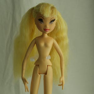 Könnte beinhalten: Puppe mit schlankem, nacktem Körper und langem, hellgelbem Haar, das zu Zöpfen frisiert ist. Die Puppe hat detaillierte Gesichtszüge, einschließlich großer Augen und eines aufgemalten Lächelns. Sie steht auf einem durchsichtigen Kunststoffständer.