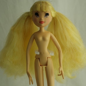 May include: A nude-colored fashion doll with long, yellow hair styled in pigtails. The doll has painted facial features, including eyes, eyebrows, and lips. It stands on a clear plastic stand, showcasing its slender physique and articulated limbs.