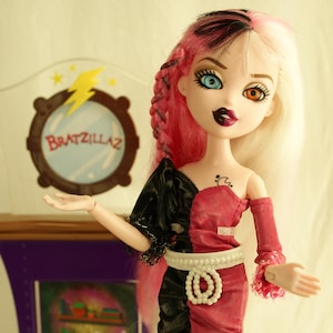 Könnte beinhalten: Eine Bratzillaz-Puppe mit rosa und weißem Haar, die ein zweifarbiges Kleid und einen Perlen-Gürtel trägt. Die Puppe hat unpassende Augen und dunklen Lippenstift. Ein lila-brauner Präsentationsständer mit dem Text "Bratzillaz" befindet sich hinter der Puppe.