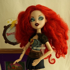 Könnte beinhalten: Bratz-Puppe mit langen, roten Haaren, trägt einen Jeansrock, ein schwarzes Oberteil und Stiefel. Die Puppe steht neben einem lila Schrank mit einem Bildschirm, der ein Bücherregal zeigt. Ein Pinsel liegt auf der Basis. Das Bratz-Logo ist sichtbar.