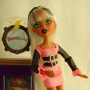 Könnte beinhalten: Eine Bratzillaz-Puppe mit langen weißen Haaren und grünen Augen, die ein rosa Kleid mit schwarzen Ärmeln und Stiefeln trägt. Die Puppe hat ein Tattoo auf dem Bein und posiert neben einem lila Ständer mit dem Bratzillaz-Logo.