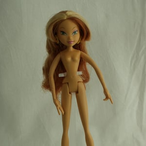 May include: A tan-skinned fashion doll with long, wavy auburn hair and green eyes. The doll is standing on a clear plastic stand. The doll has a slender build and is wearing no clothing.