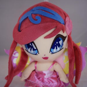 Pode incluir: Uma boneca de pelúcia com cabelo vermelho vibrante, uma faixa azul e olhos grandes azuis. A boneca veste um vestido rosa com padrão de bolinhas e apresenta asas coloridas e sobrepostas em tons de amarelo e roxo presas às costas da figura.