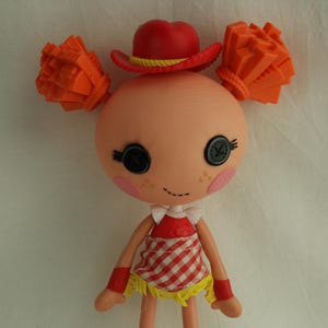 May include: A Lalaloopsy doll with a round head, button eyes, and rosy cheeks. The doll wears a red cowboy hat, orange hair puffs, a red and white checkered dress, and red boots. The doll has a stitched smile.