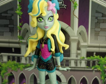 May include: A Monster High doll with yellow hair, blue accents, and green skin. The doll wears a black and pink outfit with black shorts and white stockings. The doll is standing on a base that says "Monster High".