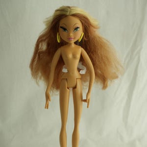 May include: A fashion doll with tan skin, long, layered auburn hair, and green eyes. The doll has yellow hoop earrings and is standing on a clear plastic base. The doll is wearing no clothing.