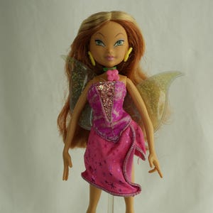 May include: A doll with long auburn hair, green eyes, and yellow earrings. The doll wears a pink dress with a star pattern and glittery wings. A pink flower is on the doll's neck.