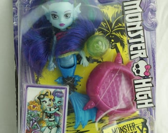 Peut inclure: Poupée Monster High Lagoona Blue dans un emballage plastique transparent. La poupée a la peau bleue, de longs cheveux bleus et une queue de sirène. L'emballage comprend un accessoire rose et une petite carte avec des illustrations.