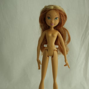 May include: A tan-skinned fashion doll with long, wavy auburn hair and green eyes. The doll is nude and stands on a clear plastic stand. The doll's arms are outstretched, and it has a slender build. The doll's face has a slight smile.