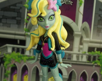 May include: A Monster High doll with pale green skin, yellow hair, and blue accents. She wears a black and pink outfit and stands on a platform with the Monster High logo. The background features a gothic-style building.