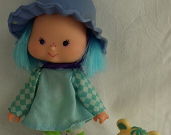 May include: A blue-haired doll with a blue hat and a checkered dress. The doll is wearing green and white striped stockings and pink shoes. The doll is holding a yellow toy dog with a blue and white checkered bib.