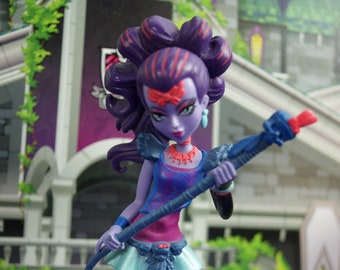 May include: A Monster High doll with purple skin, hair, and clothing. The doll is holding a blue staff with a red tip. She wears a blue and pink top, a light blue skirt, and black leggings. The doll stands on a purple base with the Monster High logo.